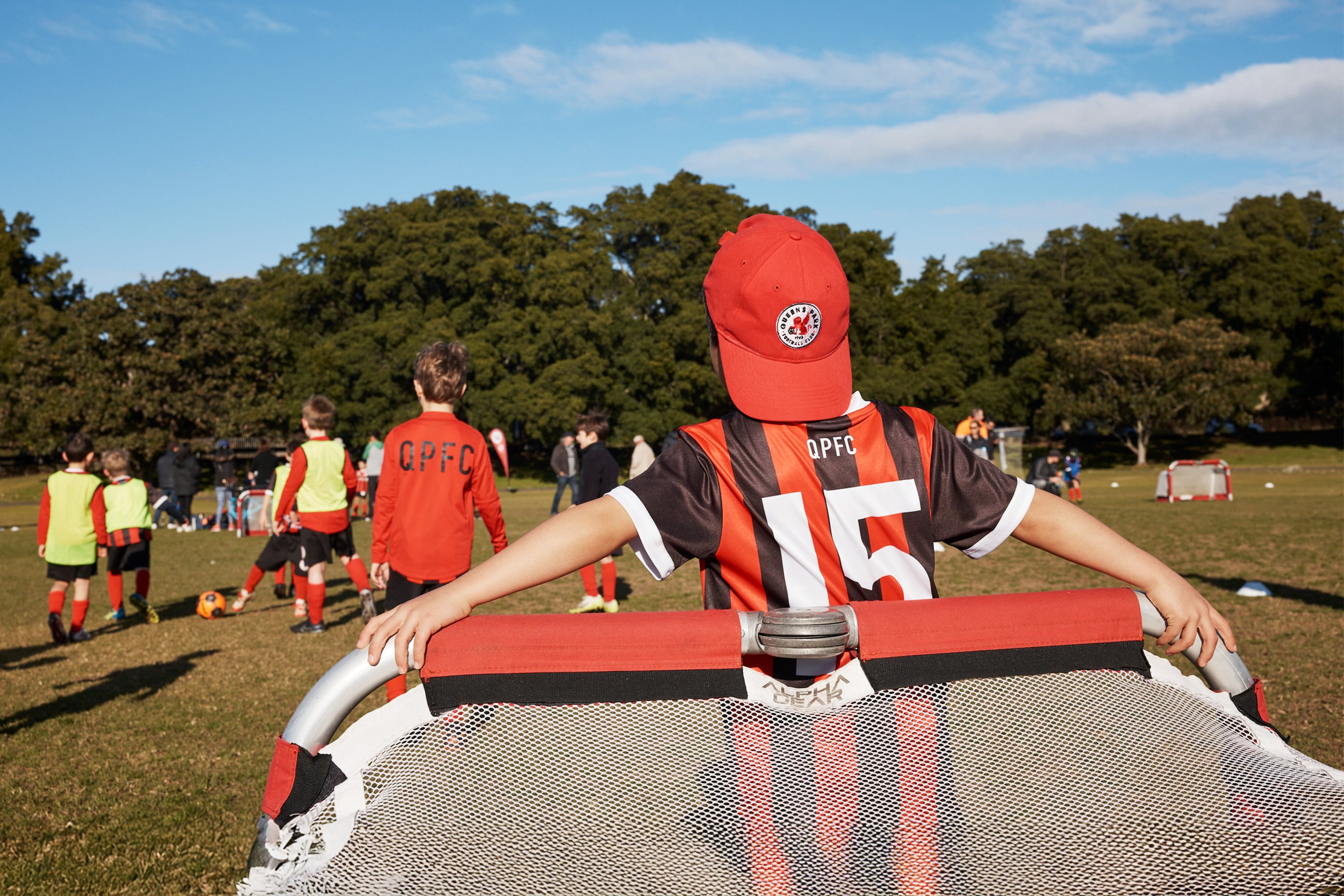 Boy standing next to football nets wearing QPFC red cap with red and black striped QPFC shirt